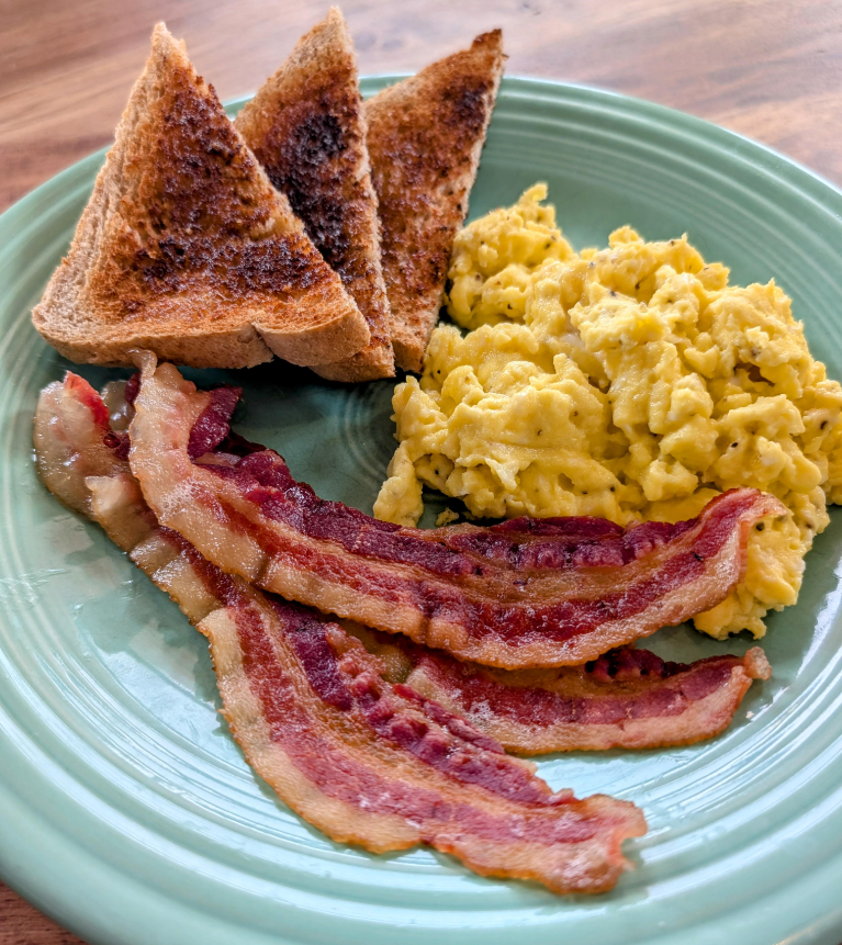 A green plate with scrambled eggs, three slices of crispy bacon, and three triangular pieces of toasted bread—classic everyday products—on a wooden table.