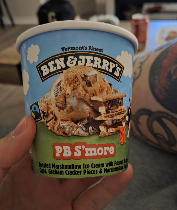 A hand holding a pint of Ben & Jerry’s PB S’more ice cream, one of those irresistible everyday products, with images of ice cream, peanut butter cups, and graham crackers on the label. A tattooed arm and blurred furniture appear in the background.