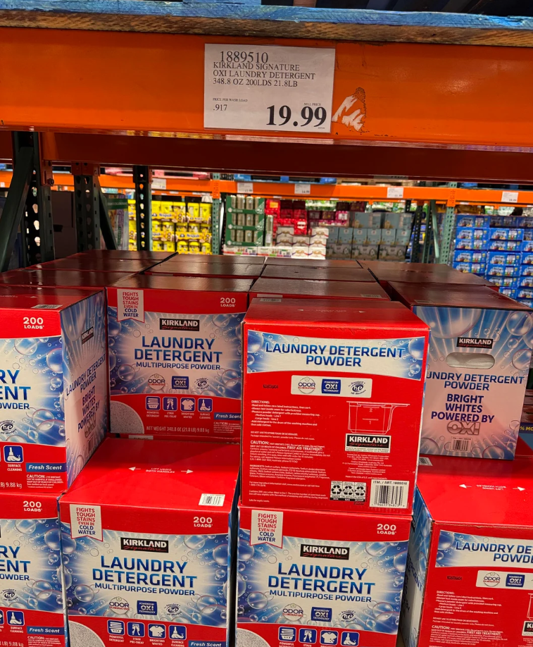 Boxes of Kirkland Signature Laundry Detergent Powder, an essential among everyday products, are stacked on a Costco shelf. A price sign above displays $19.99 for a 12.7 kg (200 load) box, with other products visible in the background.