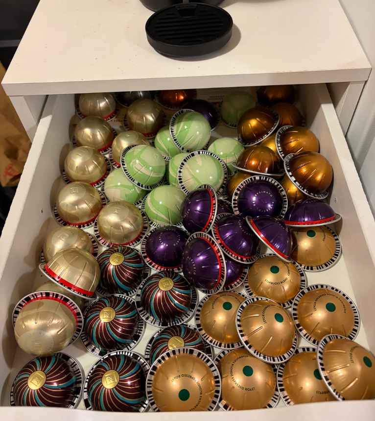 A drawer filled with various colorful coffee pods—gold, green, bronze, purple, and striped—organized in a somewhat scattered manner. An open coffee pod container lid rests on top of the drawer, highlighting the variety of everyday products inside.