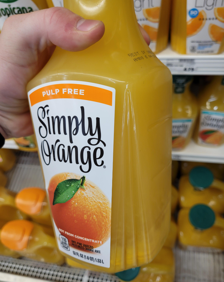 A hand holds a bottle of Simply Orange pulp-free orange juice—one of those everyday products— in front of a store shelf stocked with more bottles of the same brand.