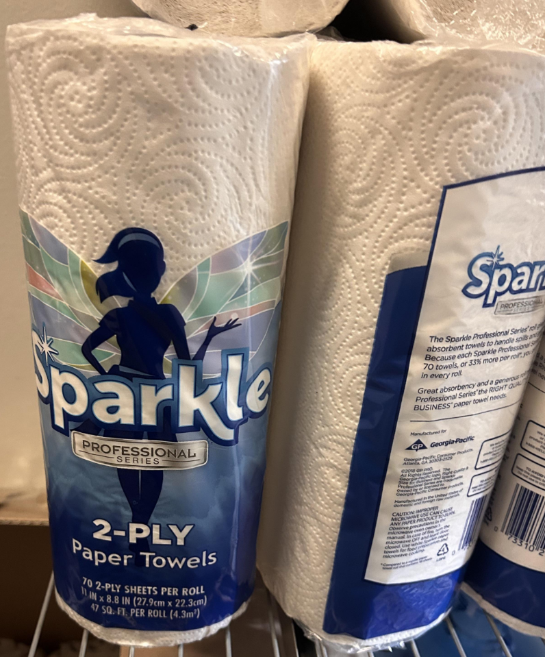 A roll of Sparkle Professional Series 2-ply paper towels, an essential among everyday products, is shown on a shelf, with the front label featuring a blue silhouette of a woman and colorful accents, next to another roll displaying the product information.
