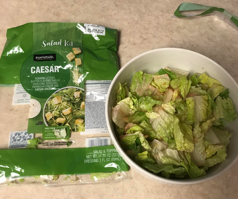 An opened package of Marketside Caesar Salad Kit, one of your favorite everyday products, sits on a countertop next to a bowl filled with prepared Caesar salad made from the kit.