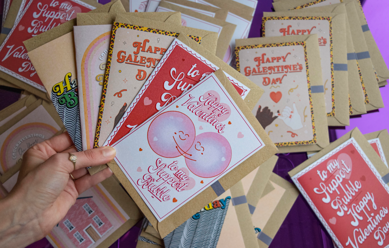 A hand holds a fan of colorful Valentine and Galentine’s Day cards—everyday products with cheerful designs and messages. Additional cards are spread out on a purple surface in the background.