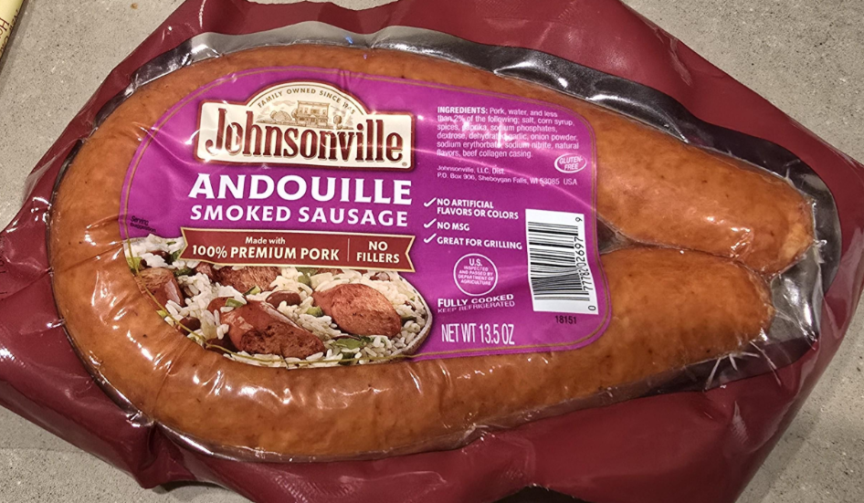 A package of Johnsonville Andouille Smoked Sausage made with 100% premium pork, shown on a countertop. The sausage is horseshoe-shaped and vacuum sealed in clear plastic with a purple label.