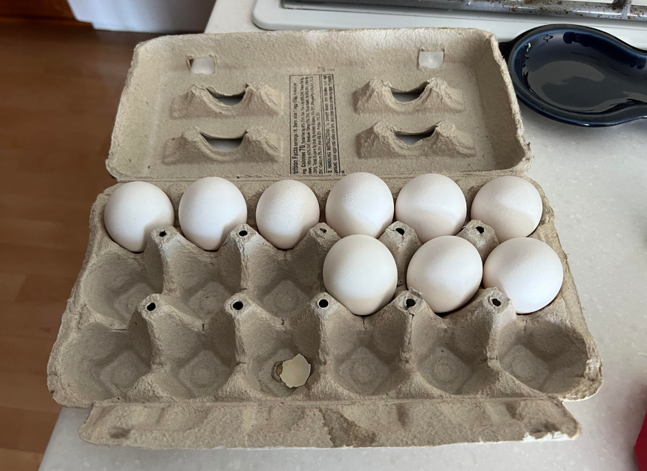 A cardboard egg carton, one of many everyday products, sits open on a counter holding ten white eggs in two rows, with two spaces empty in the front row.