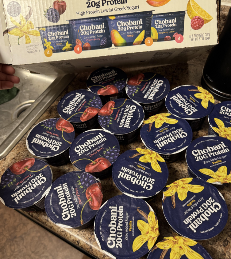 Several containers of Chobani 20g Protein Greek Yogurt in blueberry, vanilla, and strawberry flavors are spread out on a kitchen counter—everyday products with the packaging box visible in the background.