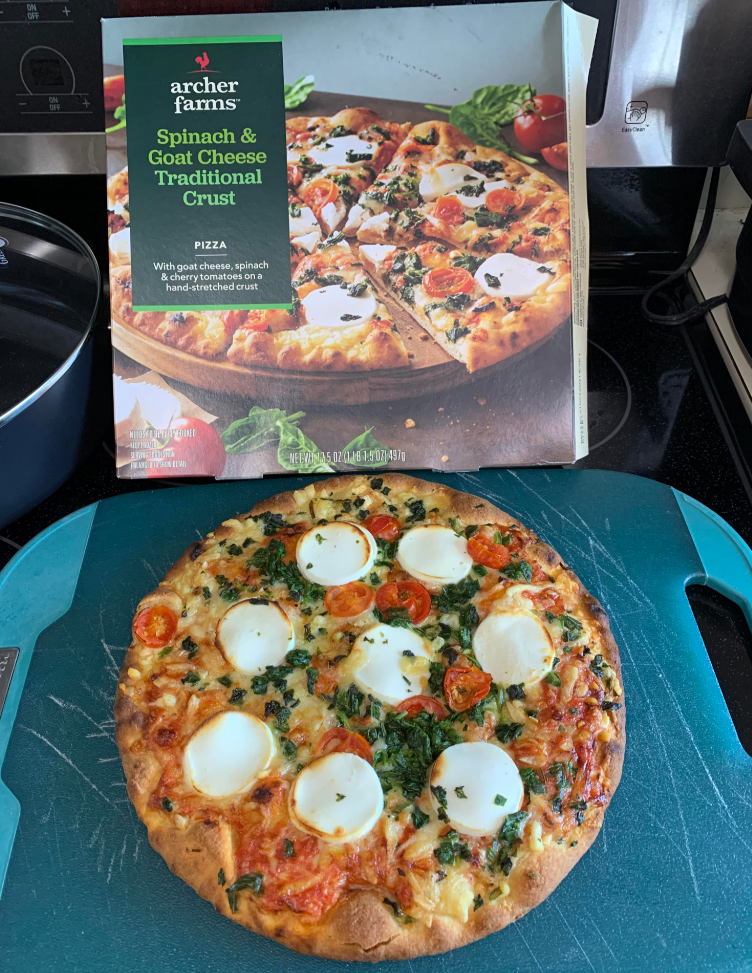 A baked Archer Farms spinach and goat cheese pizza, one of those delicious everyday products, sits on a cutting board in front of its box, featuring a tempting image of spinach, tomatoes, and goat cheese rounds on a traditional crust.