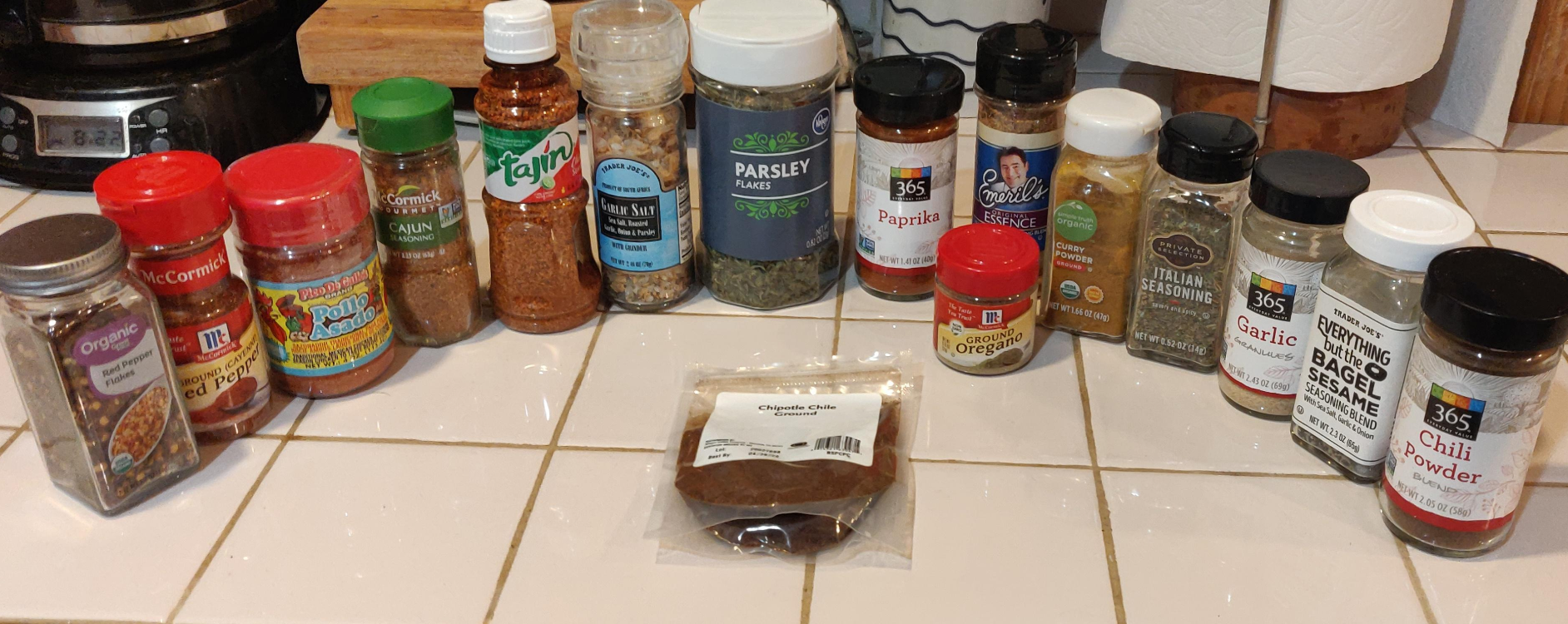 Sixteen everyday products, including oregano, cayenne, paprika, garlic powder, parsley flakes, and chili powder, are arranged in a semicircle on a kitchen counter with a sealed bag of chipotle chili in the center.