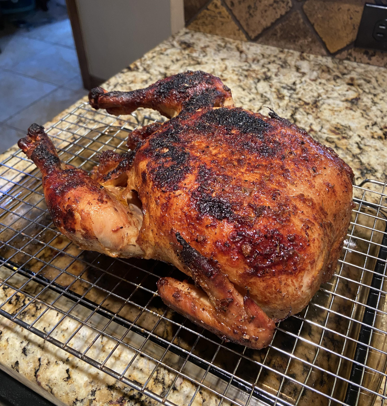A whole roasted chicken with crispy, browned skin rests on a metal cooling rack—one of those essential everyday products—over a granite countertop in a kitchen.