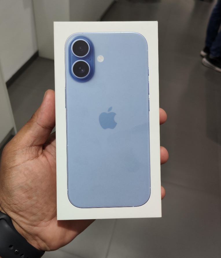 A hand holds a sealed box of a blue iPhone with dual cameras, featuring the Apple logo on the back, in a store setting with a gray floor—highlighting how products are getting more expensive because of tariffs.