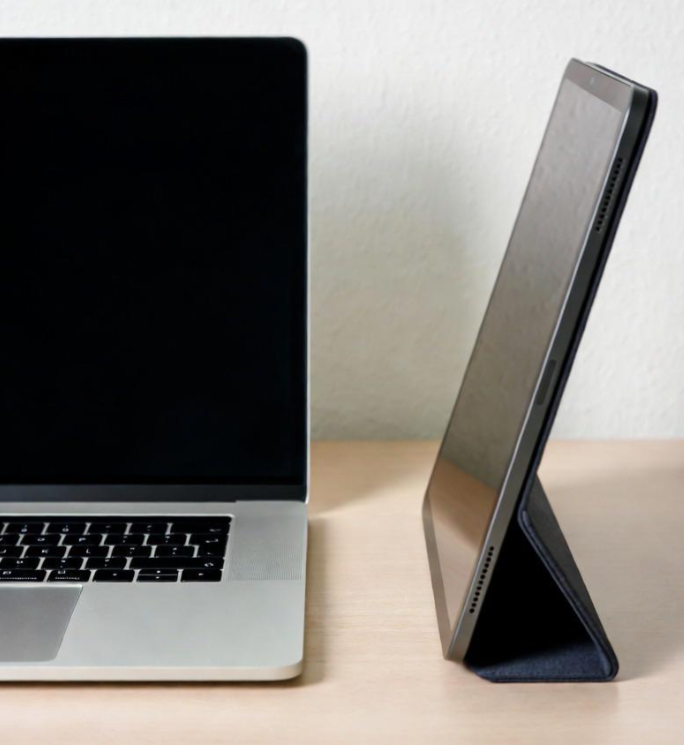 A laptop and a tablet in a case, both screens off, sit side by side on a light wooden desk against a plain white wall—reminders of how products are getting more expensive because of tariffs.