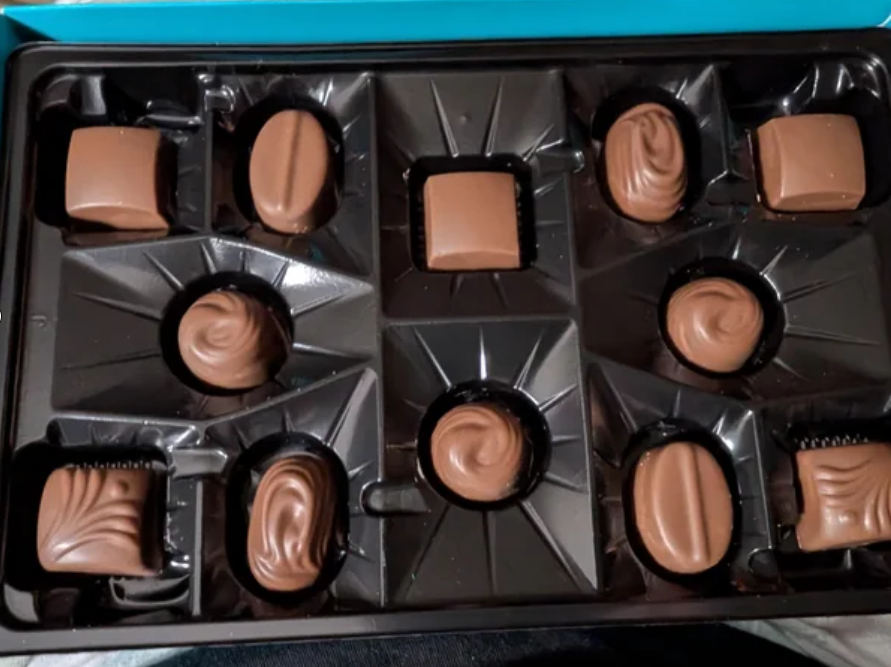 A box of assorted chocolates with various shapes and designs sits open, with several empty spaces where chocolates have been removed.