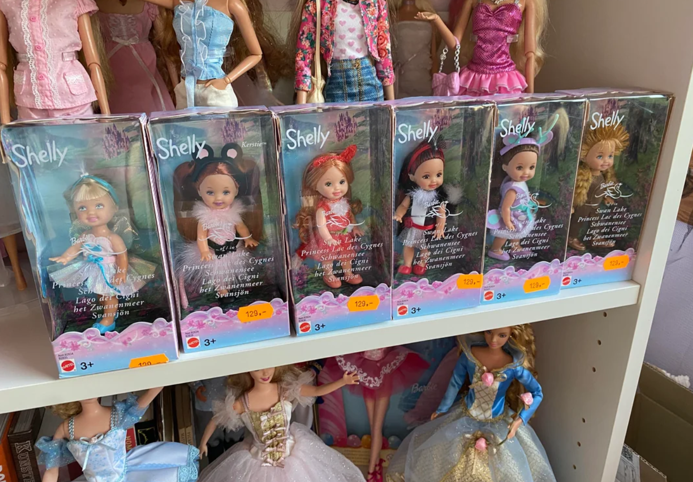 A row of five boxed Shelly dolls in fairy-tale costumes is displayed on a shelf, reminding collectors that beloved products are getting more expensive because of tariffs, with more dressed dolls standing and sitting on shelves above and below.