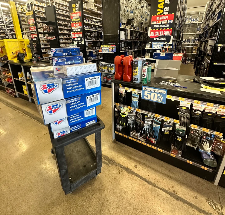 A cart stacked with blue Carquest oil filters stands near a counter displaying gloves, car fluids, and a "Clearance Event 50% Off" sign—welcome savings as products are getting more expensive because of tariffs in this auto parts store aisle.