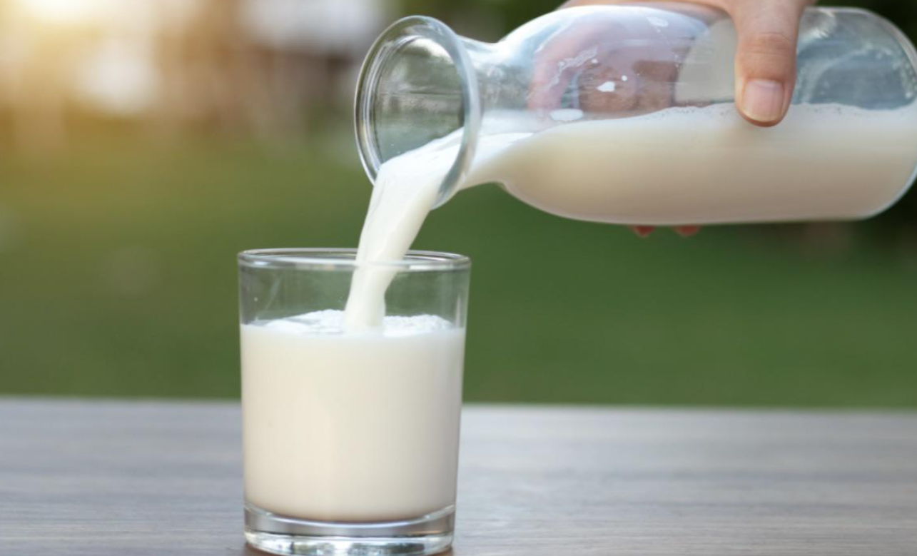 A hand pours milk from a glass jug into a clear glass on a wooden surface, with a blurred green outdoor background—a reminder of how products are getting more expensive because of tariffs.