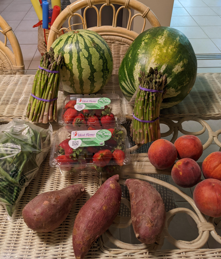 A wicker table displays watermelons, asparagus bundles, strawberries in plastic containers, green beans in a bag, three sweet potatoes, and four peaches—products getting more expensive because of tariffs. Wicker chairs sit around the table.