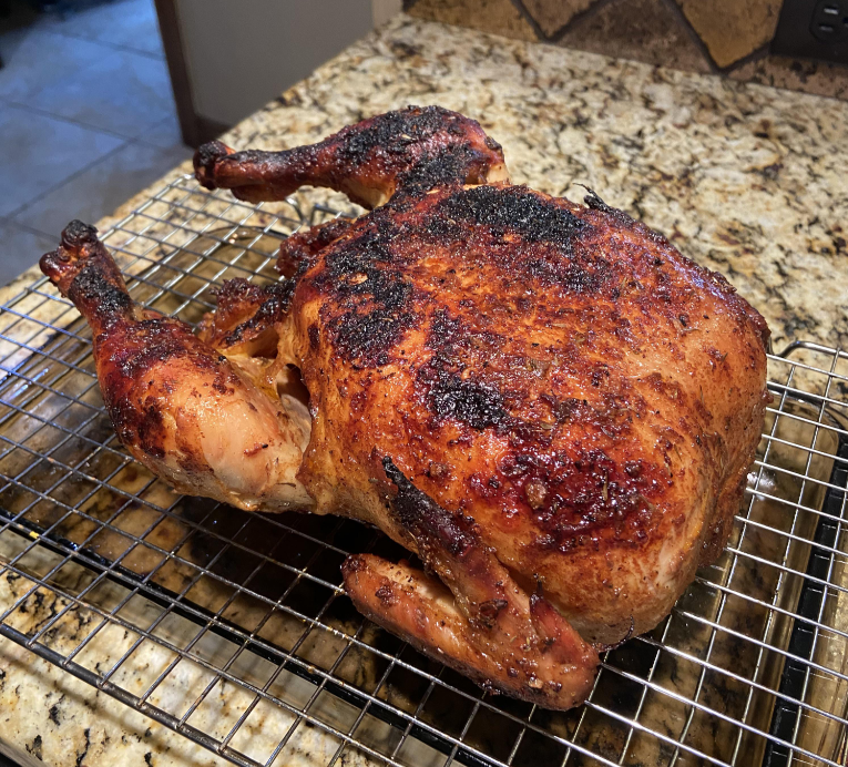 A whole roasted chicken with crispy, browned skin rests on a wire rack atop a granite countertop—a simple pleasure that feels even more special with products getting more expensive because of tariffs.