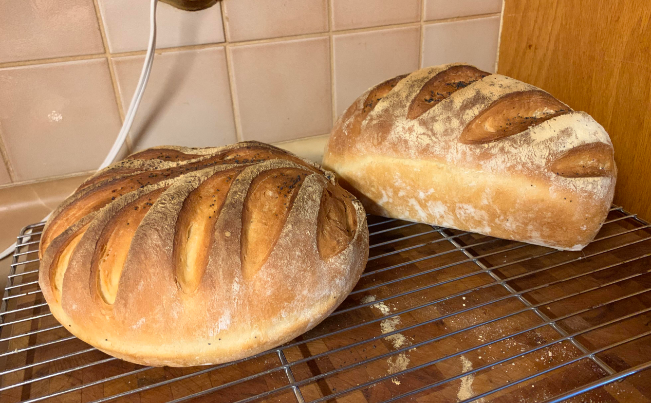 Two round loaves of homemade bread with golden-brown crusts and deep slashes on top, cooling on a wire rack in a cozy kitchen—a comforting sight as products are getting more expensive because of tariffs.