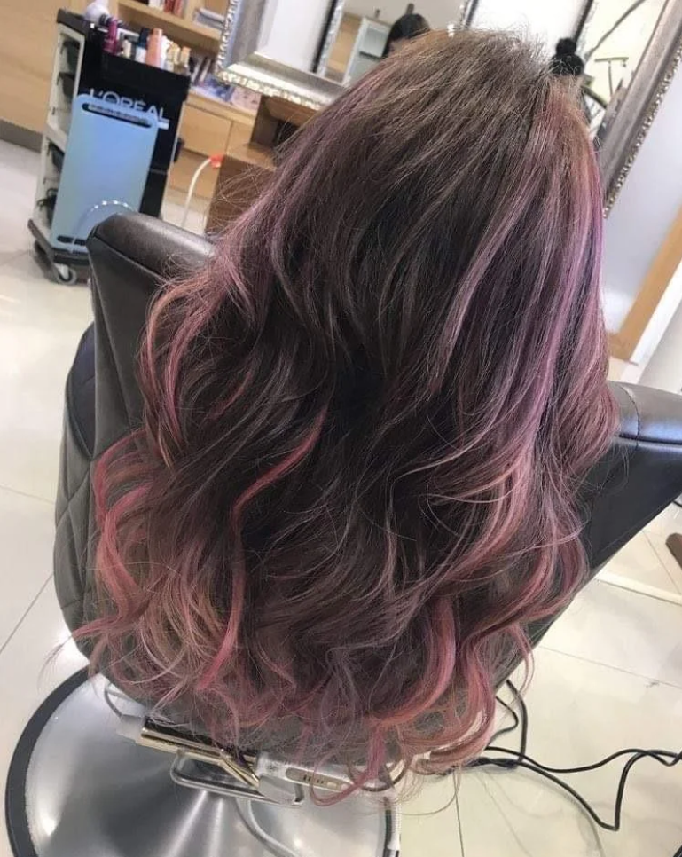 A person with long, wavy brown hair featuring soft, pastel pink highlights is seated in a salon chair, surrounded by salon equipment and mirrors—perhaps reflecting on products getting more expensive because of tariffs.
