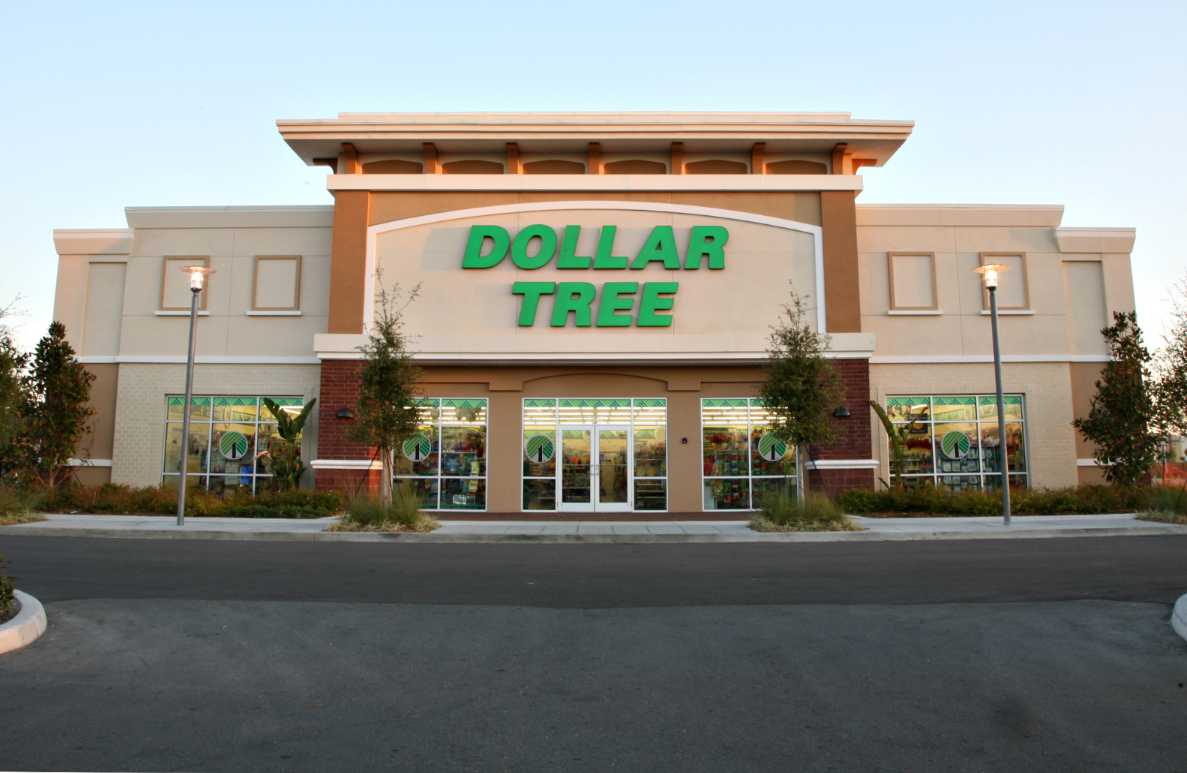 A Dollar Tree store with a beige and brown exterior, large Dollar Tree green sign above the entrance, glass windows displaying products, and a small parking lot in front.