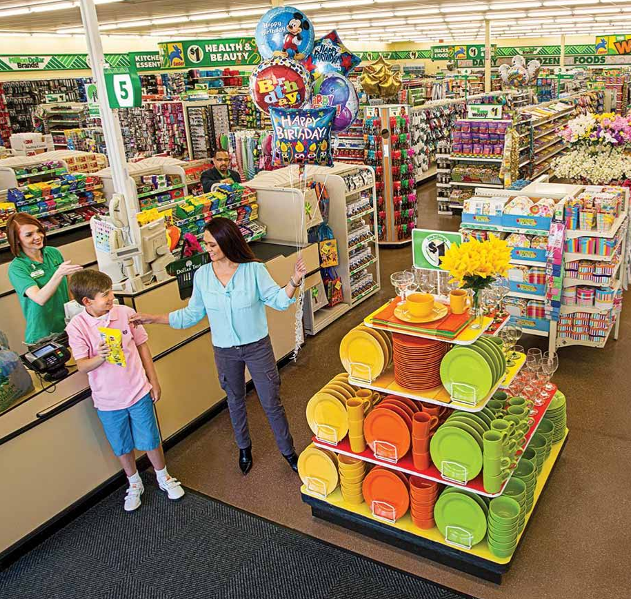 A woman and a boy stand near a colorful display of plates and cups in a Dollar Tree. The boy holds candy as a cashier smiles at them. The bright store is filled with various items, including birthday balloons.