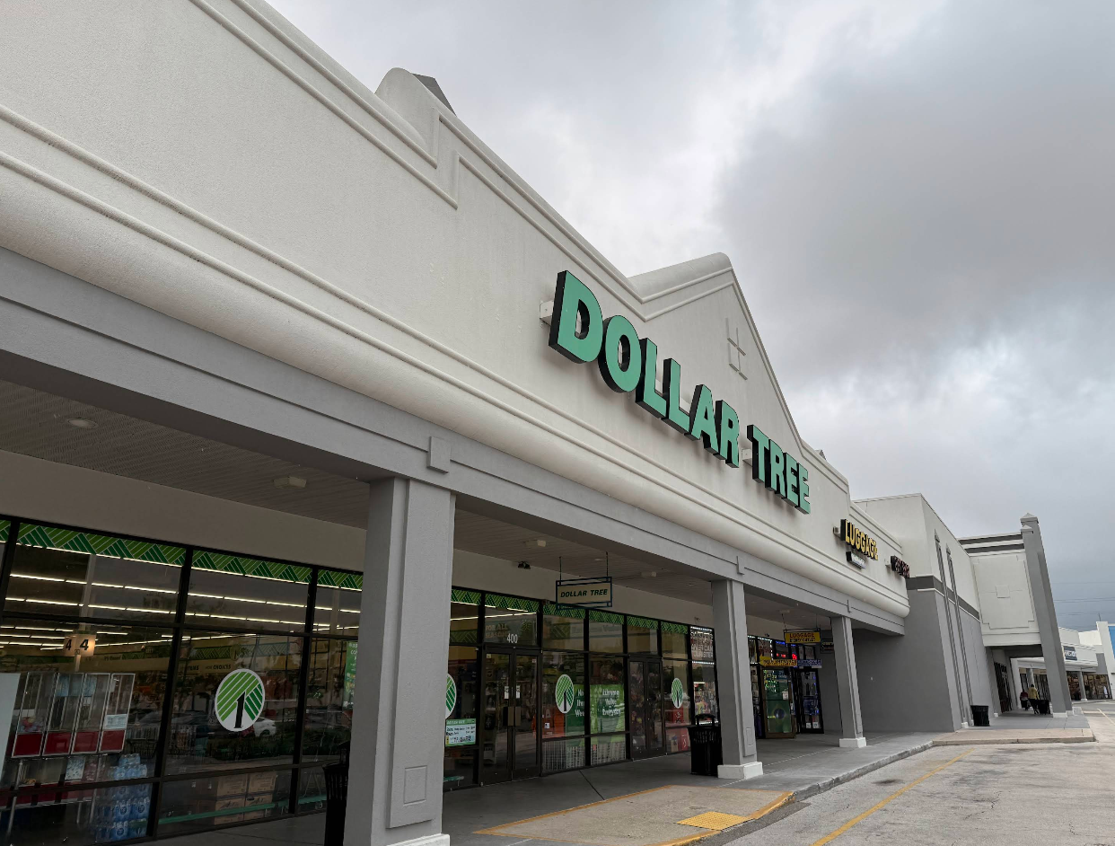 Exterior view of a Dollar Tree store in a shopping plaza, with the iconic large green Dollar Tree signage above the entrance and overcast skies in the background. Storefront windows display items and the parking lot is visible.