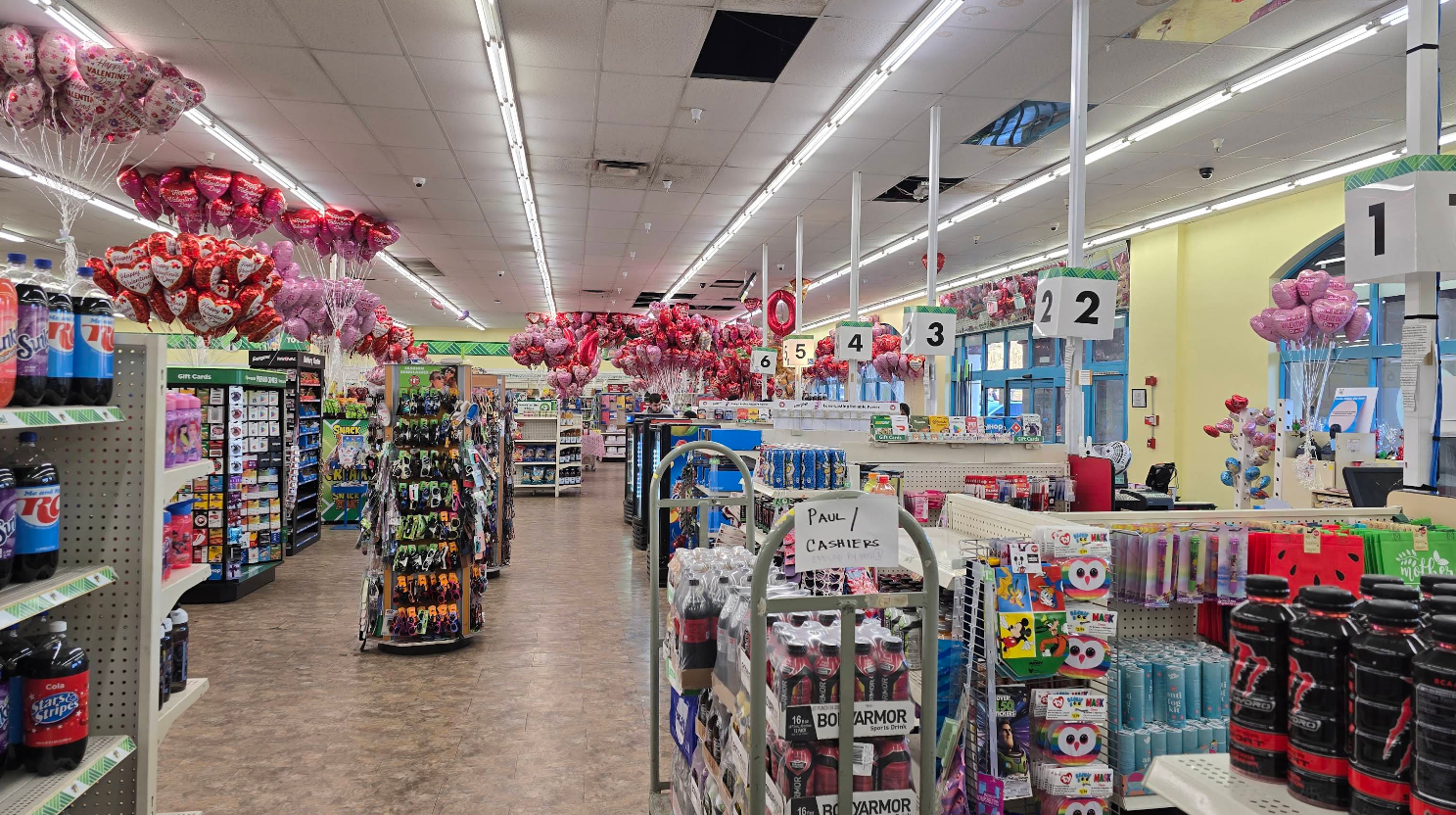 A brightly lit and organized Dollar Tree grocery store is decorated for Valentine's Day with pink and red heart-shaped balloons. Numbered checkout lanes are visible, along with shelves stocked with drinks and snacks.