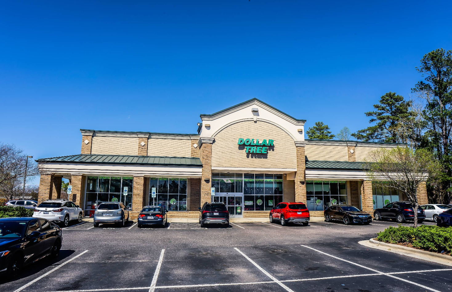 A Dollar Tree store with a beige exterior, green roof, and large glass windows sits under a clear blue sky. Several cars are parked in the lot on this sunny day, with trees framing the Dollar Tree location in the background.