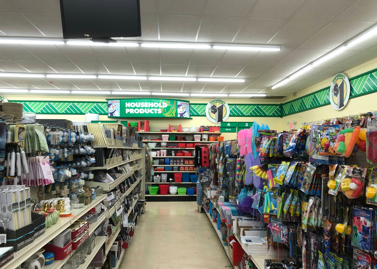 A brightly lit Dollar Tree aisle displays an array of household products on shelves, including cleaning supplies, kitchenware, bins, and toys. A green sign above reads “Household Products” with a dollar sign symbol.