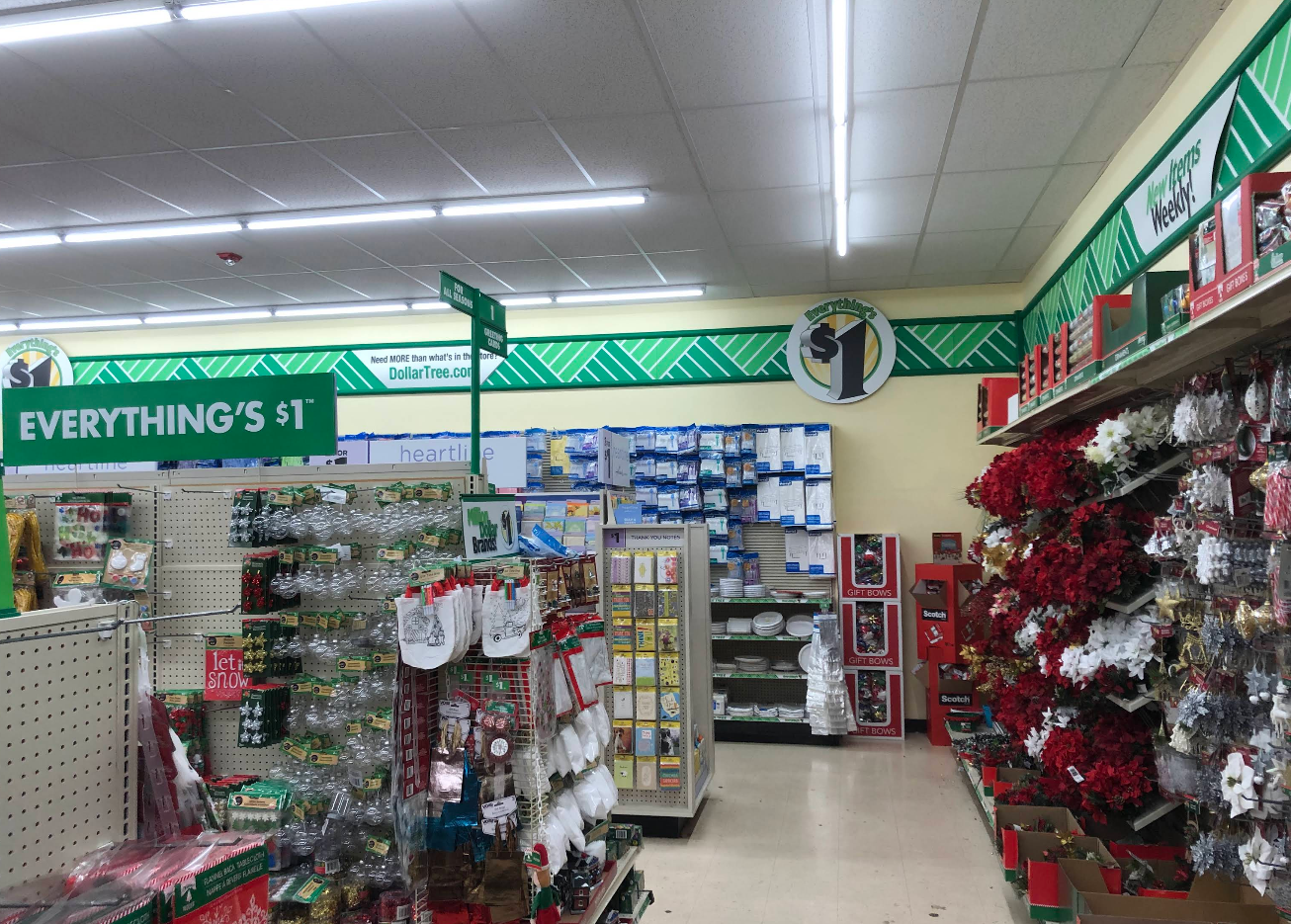 Aisle in a Dollar Tree store decorated for the holidays, with shelves displaying Christmas ornaments, flowers, household goods, and a large sign reading "Everything's $1" above the aisle.