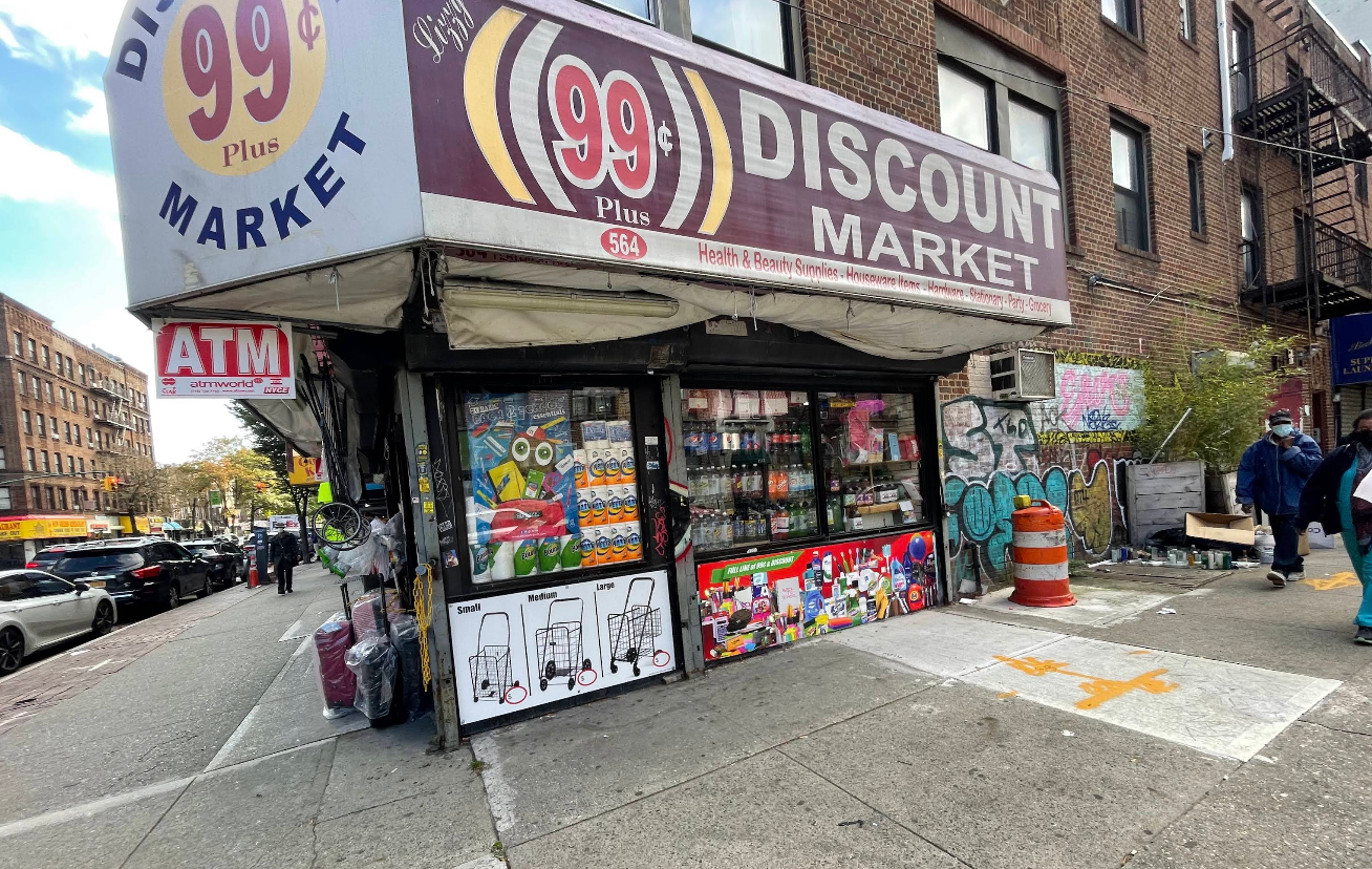 A 99¢ Plus Discount Market, reminiscent of a Dollar Tree, sits on a city corner with goods displayed outside, a red ATM sign, colorful posters in the window, and graffiti on the wall. People pass by as cars line the busy street.