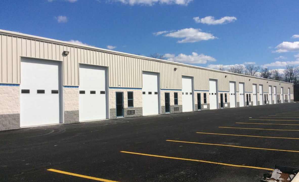 A row of industrial warehouse units, including a Dollar Tree, with large white garage doors and windows, set along a recently paved parking lot with yellow lines on a sunny day with blue sky and scattered clouds.