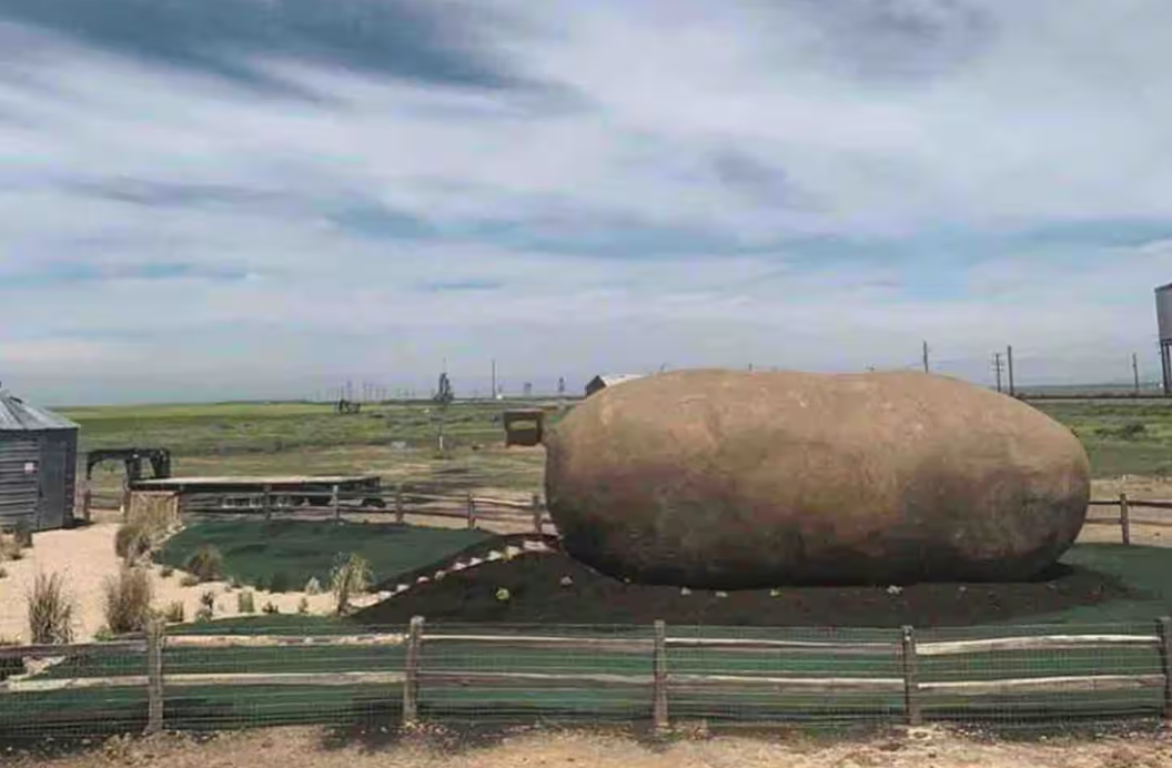A giant potato-shaped structure sits in a fenced area on a grassy field under a partly cloudy sky, with farm buildings and equipment in the background—one of the coolest Airbnbs for a quirky countryside getaway.