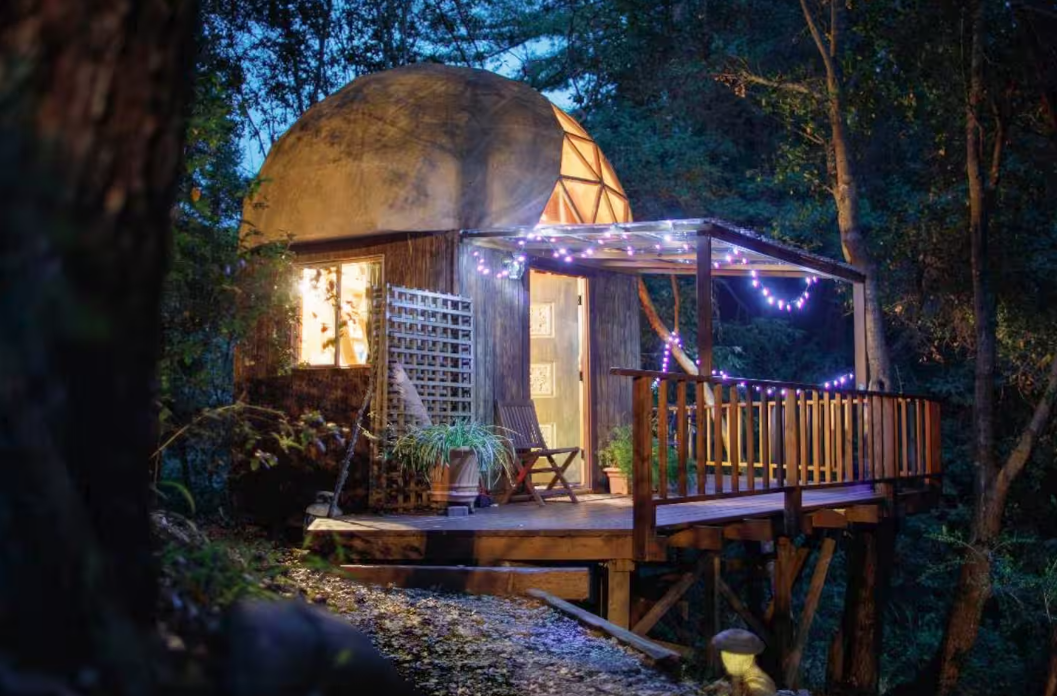 A cozy dome-shaped treehouse with warm lights and string lights on its porch sits elevated among dense trees at dusk, creating a peaceful, inviting atmosphere in the forest—truly one of the coolest Airbnbs to experience nature.
