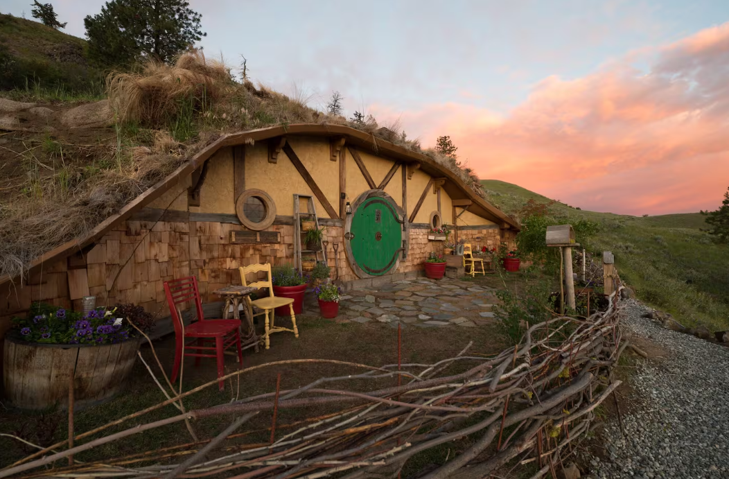 A cozy, hobbit-style house built into a grassy hillside with a round green door, rustic wood details, colorful chairs, and potted plants—a perfect escape under a pink sunset sky and one of the coolest Airbnbs you’ll find.