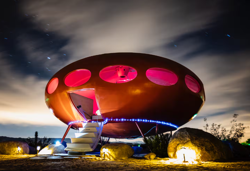 A round, UFO-shaped house—one of the coolest Airbnbs—features circular pink-lit windows, perched on rocks beneath a cloudy night sky, with steps leading to an open door and blue lights outlining its edge.