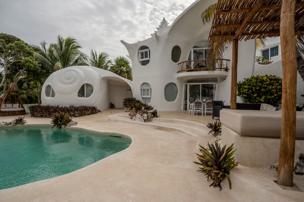 A unique white house with curved walls and seashell-like windows stands beside a pool, surrounded by tropical plants and palm trees under a partly cloudy sky—one of the coolest Airbnbs for an unforgettable escape.