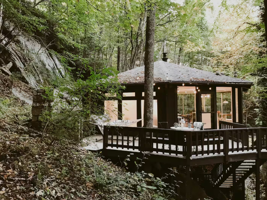 A cozy cabin with large windows and a deck sits elevated among dense, green trees in a forest—one of the coolest Airbnbs—illuminated warmly from within and surrounded by lush foliage and natural scenery.