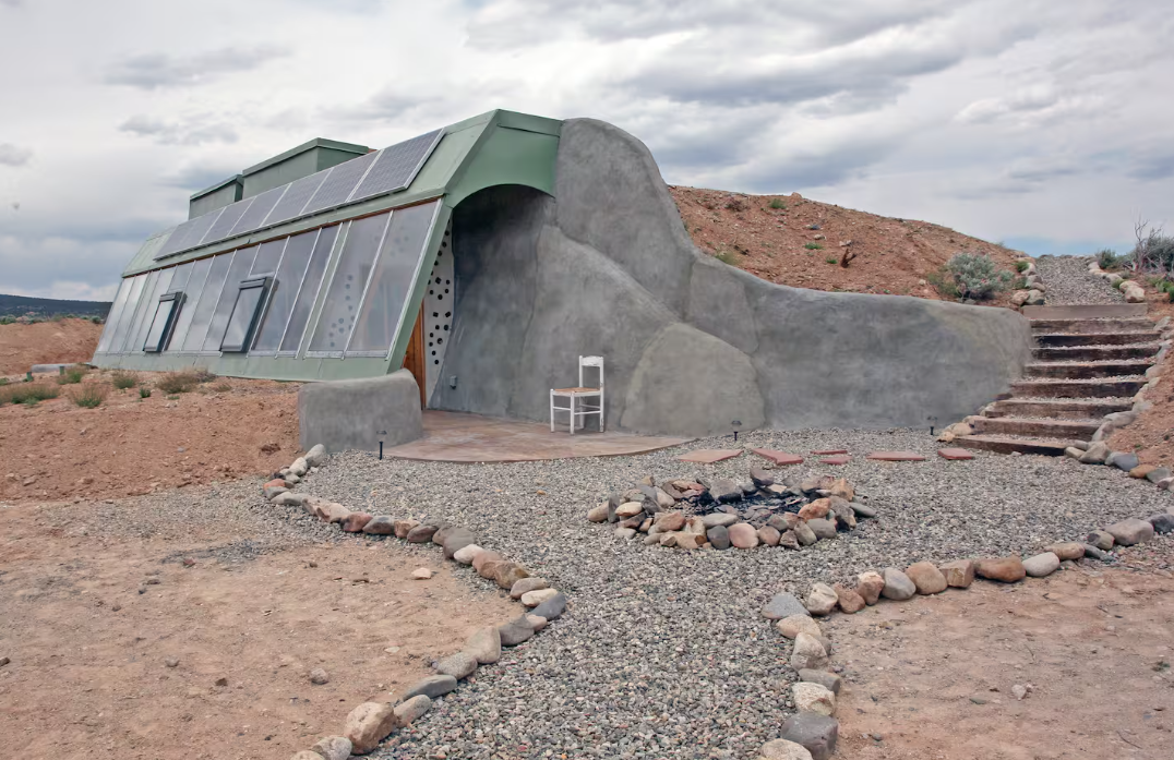 An earth-sheltered house with large south-facing windows and rooftop solar panels is built into a sandy hill—one of the coolest Airbnbs. A white chair sits on the patio by a stone-lined fire pit, curved gravel paths, and steps up the slope.