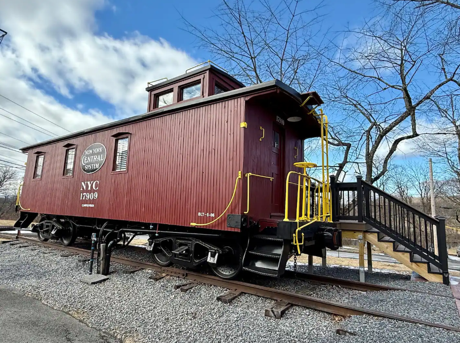 A vintage red New York Central caboose train car, featured among the coolest Airbnbs, sits on short tracks with yellow railings and a wooden staircase leading to its door, set outdoors under a partly cloudy sky.