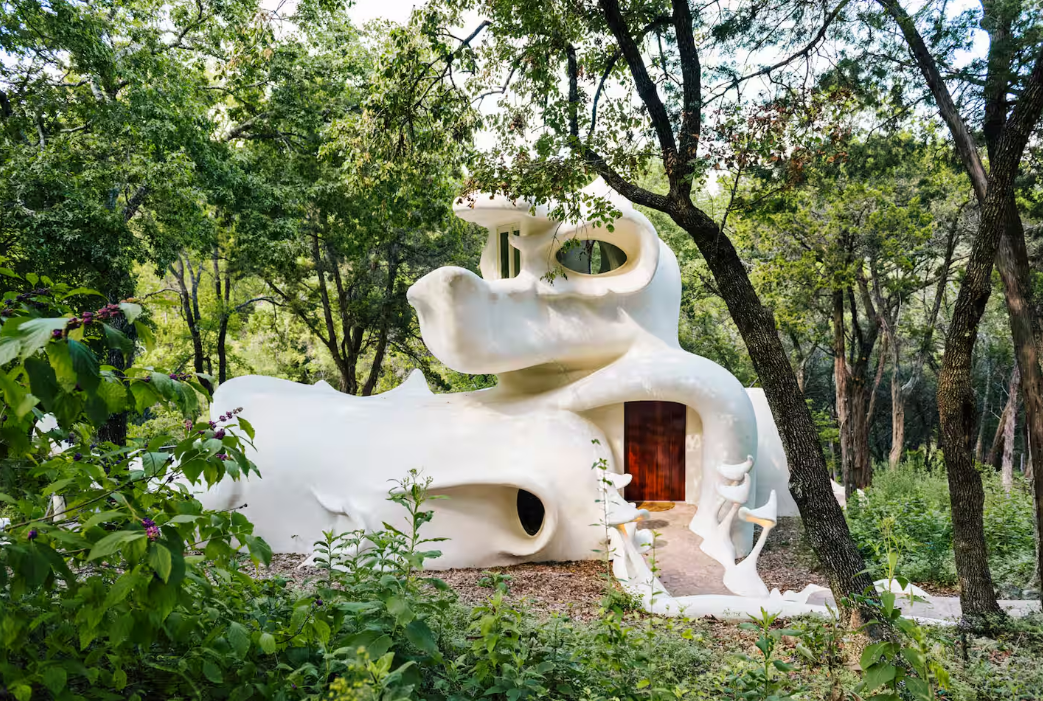 A whimsical white house with organic, curving shapes and large, rounded windows sits surrounded by lush green trees, blending into the forested landscape—one of the coolest Airbnbs for a magical, nature-inspired retreat.