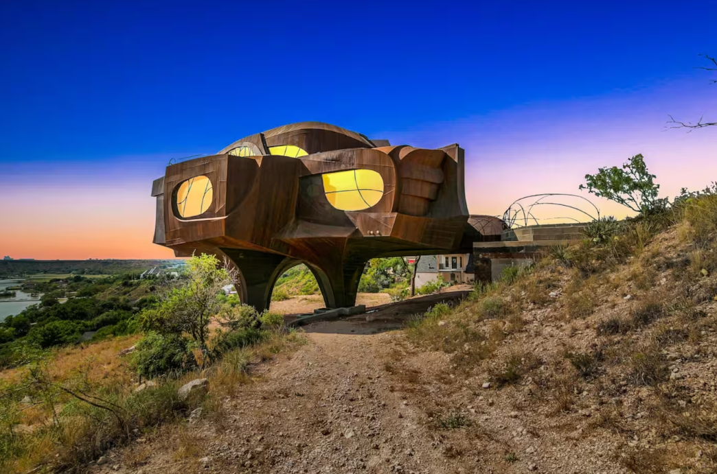A futuristic house with round windows and a metallic exterior stands elevated on stilts above rocky terrain, set against a vibrant blue and orange sunset—one of the coolest Airbnbs for an unforgettable stay.