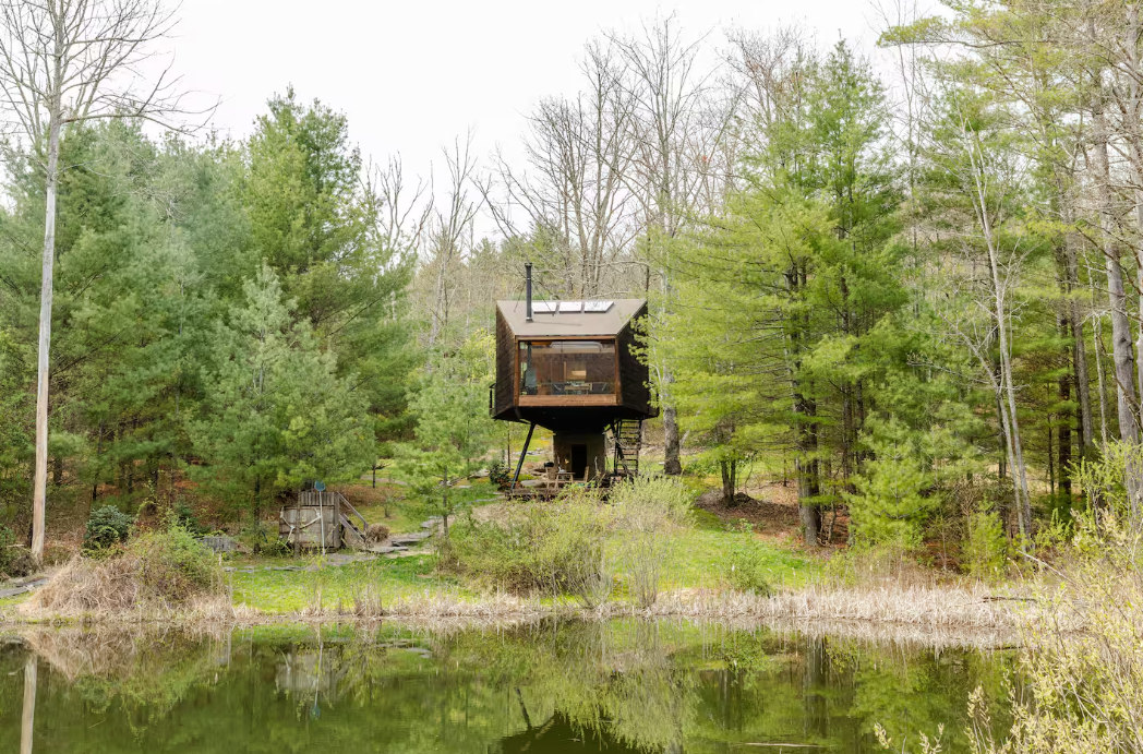 A small, modern treehouse with large windows stands elevated among tall pine trees, facing a calm pond—one of the coolest Airbnbs for enjoying serene views and nature on an early spring day.