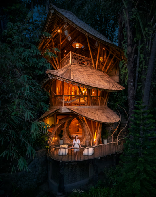 A multi-level bamboo treehouse glows warmly at dusk, surrounded by lush greenery. Among the coolest Airbnbs, a person in white relaxes on the lower terrace’s cozy outdoor seating area illuminated by soft lights.