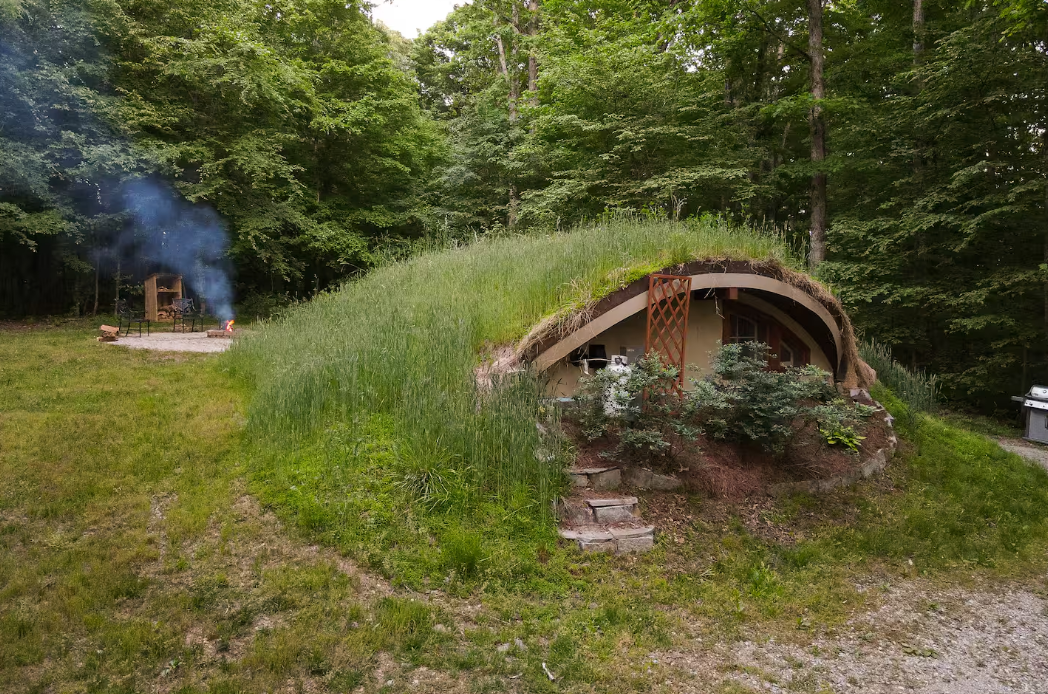 A round, earth-covered hobbit house blends into a grassy hillside surrounded by trees, with a stone path leading to its wooden door—one of the coolest Airbnbs for a magical stay. Smoke rises from a cozy fire pit area in the background.