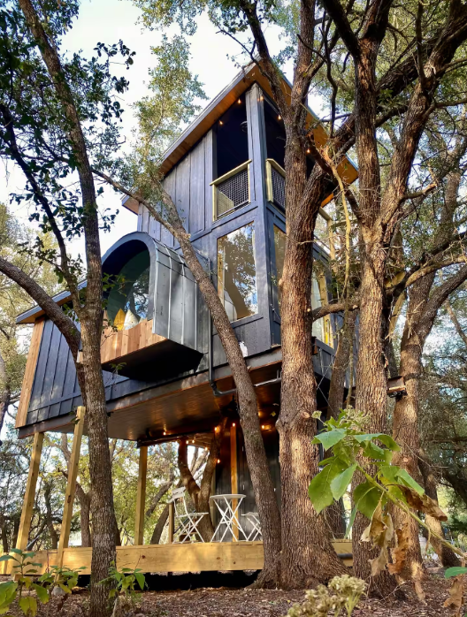 A modern two-story treehouse with large windows and a small outdoor deck sits elevated among tall trees. Often featured among the coolest Airbnbs, its deck offers two white chairs and a table, surrounded by leafy branches.