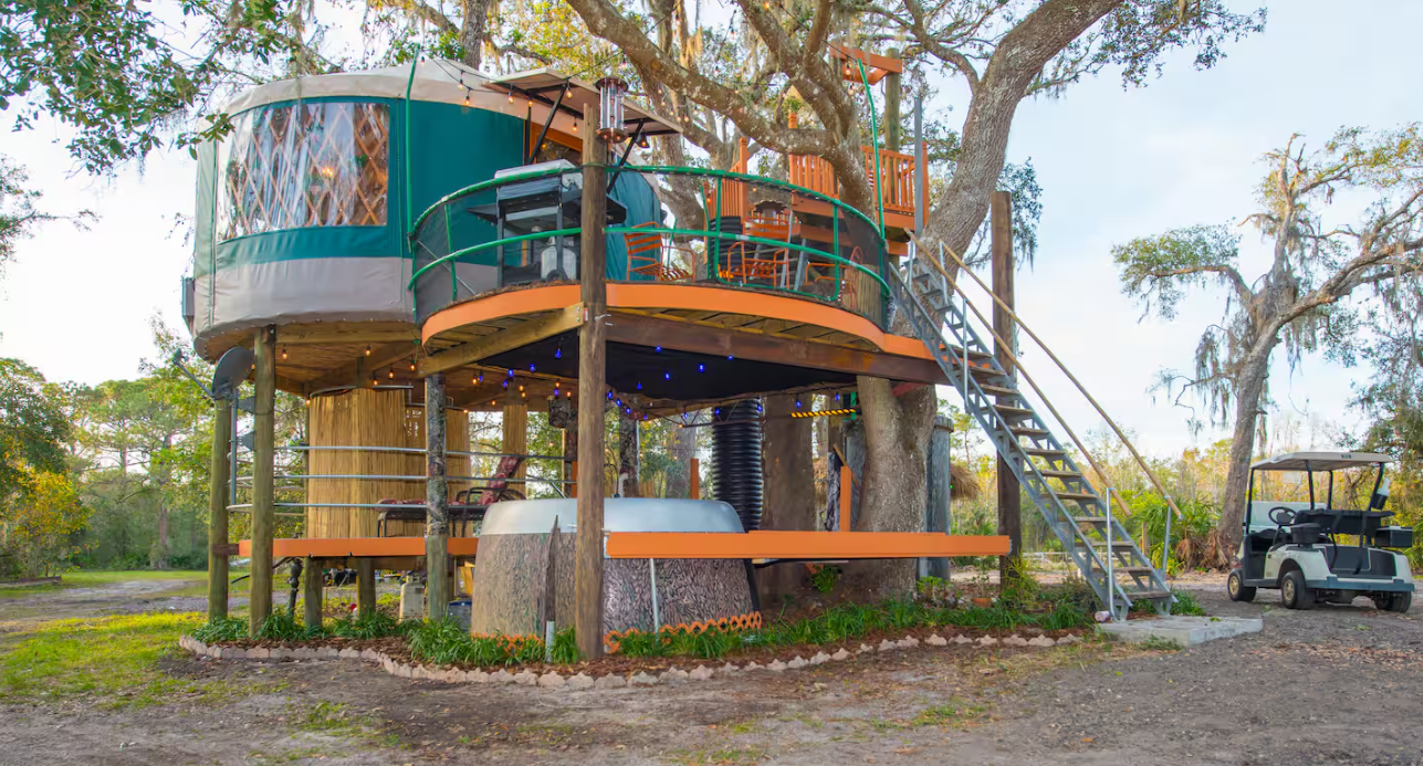 A modern treehouse with circular design stands elevated among trees, featuring two levels with railings, outdoor seating, a hot tub below, and a metal staircase. One of the coolest Airbnbs—plus, there’s a golf cart parked nearby on grassy ground.