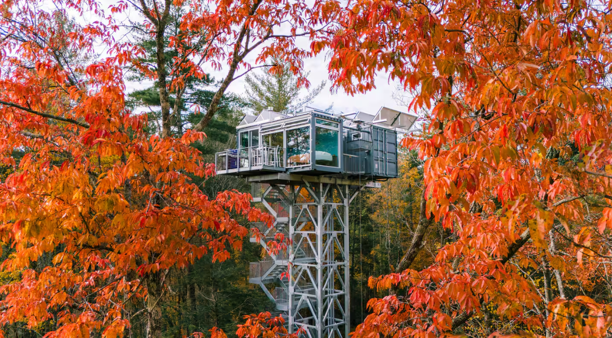 A modern glass and metal treehouse stands elevated on a tall steel structure among vibrant orange and red autumn trees, with a spiral staircase leading up to one of the coolest Airbnbs you'll ever find.