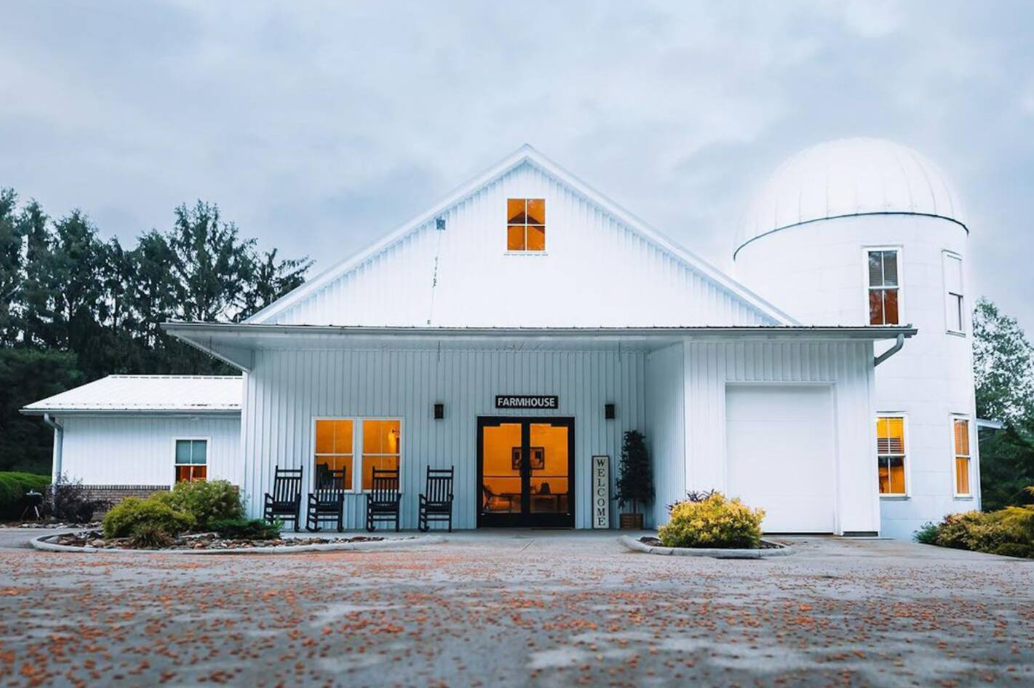 A modern white farmhouse, one of the coolest Airbnbs, with glowing windows, rocking chairs on the porch, and a round silo on the right, set against a cloudy sky and surrounded by greenery and shrubs.